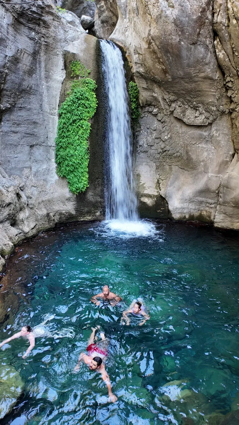 Der hohe Wasserfall im Sapadere Canyon, der zwischen Efeu in ein grünes Becken fällt, in dem mehrere Menschen schwimmen